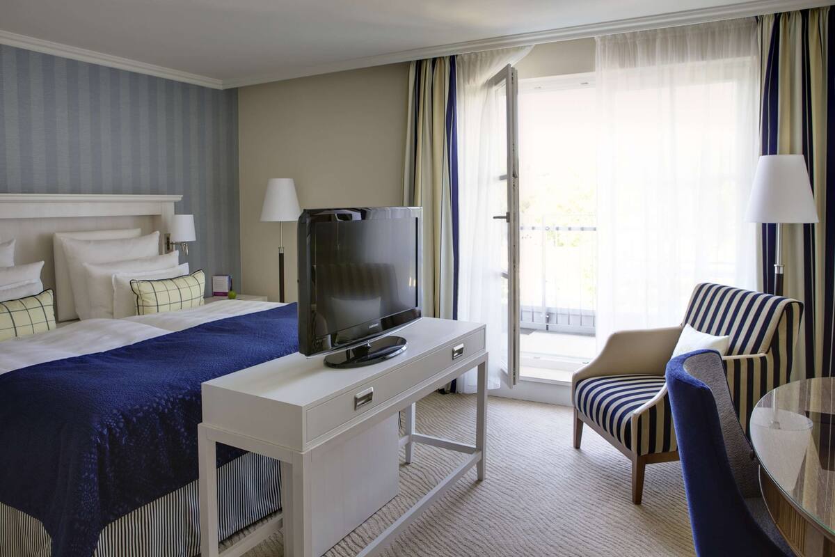 A hotel room with a bed, a TV on a white desk, a striped armchair, and a glass table.