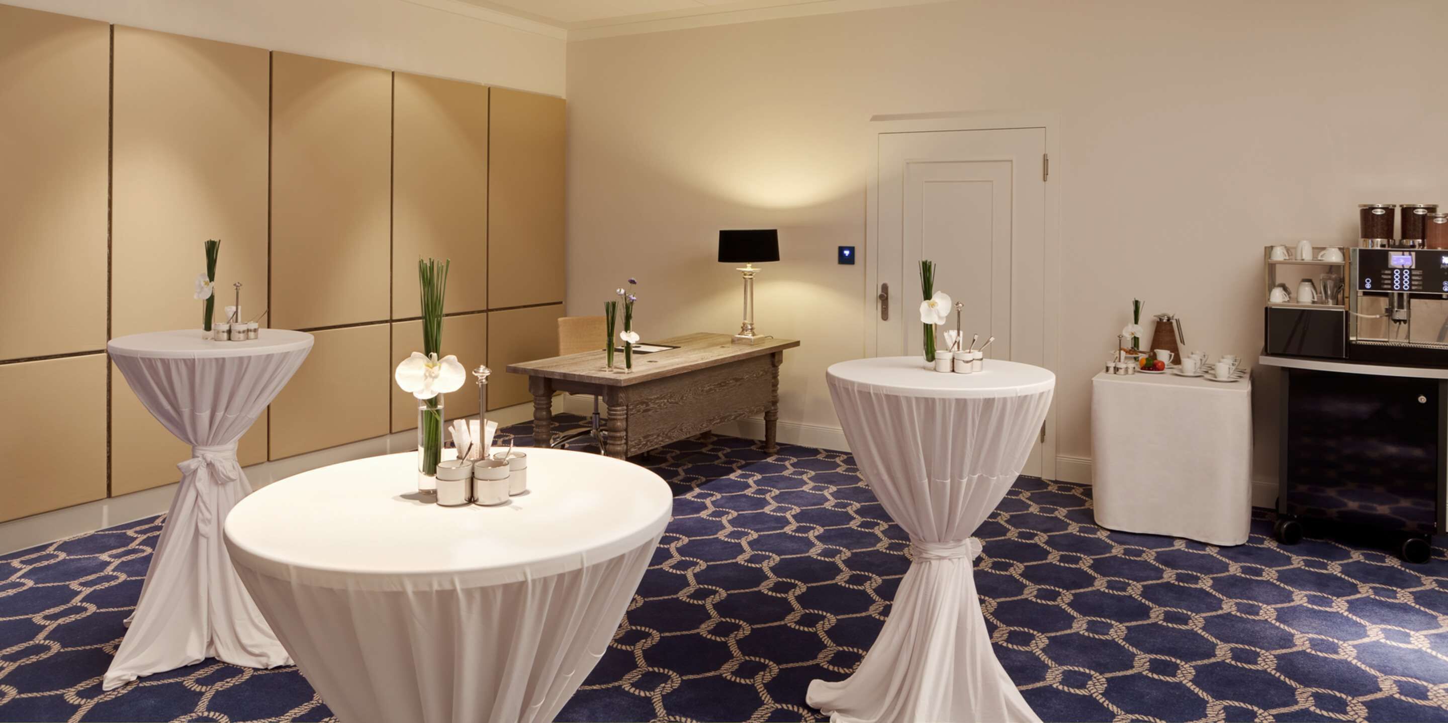 Habitación de hotel con dos mesas redondas cubiertas con manteles blancos, cada una con un jarrón de flores blancas y una pequeña vela. Se observa un minibar con distintos artículos, una cafetera y un escritorio con una lámpara. La habitación tiene una alfombra estampada y una puerta al fondo.