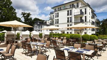 Un espace extérieur aménagé pour les repas, avec des tables, des chaises, des parasols et un bâtiment en arrière-plan.