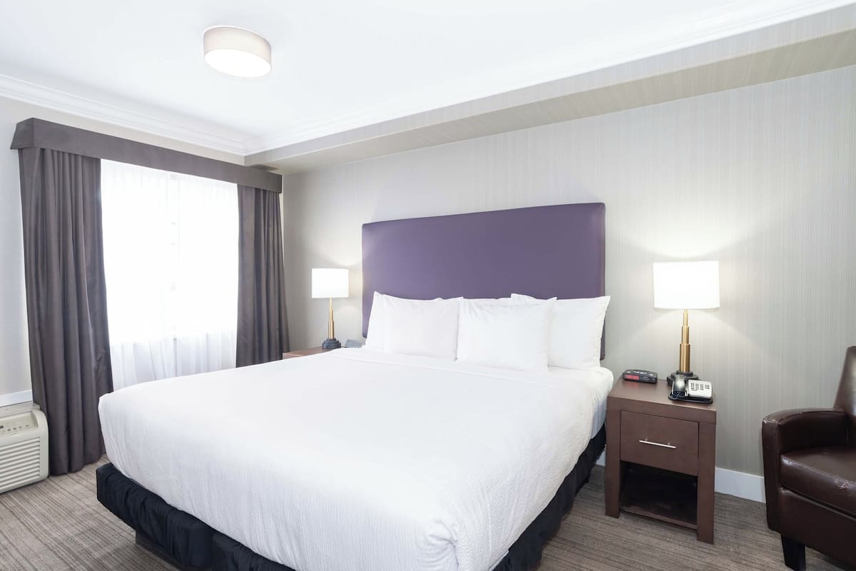 Ein Hotelzimmer mit einem großen Bett, zwei Nachttischen mit Lampen, einem Sessel und einem Fenster mit Vorhängen.