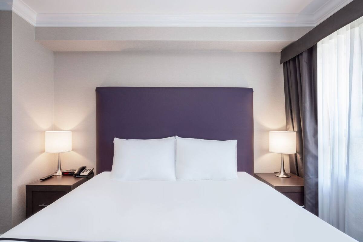 Ein Hotelzimmer mit einem großen Bett, zwei Nachttischen, einem Telefon und einem Fenster mit Vorhängen.
