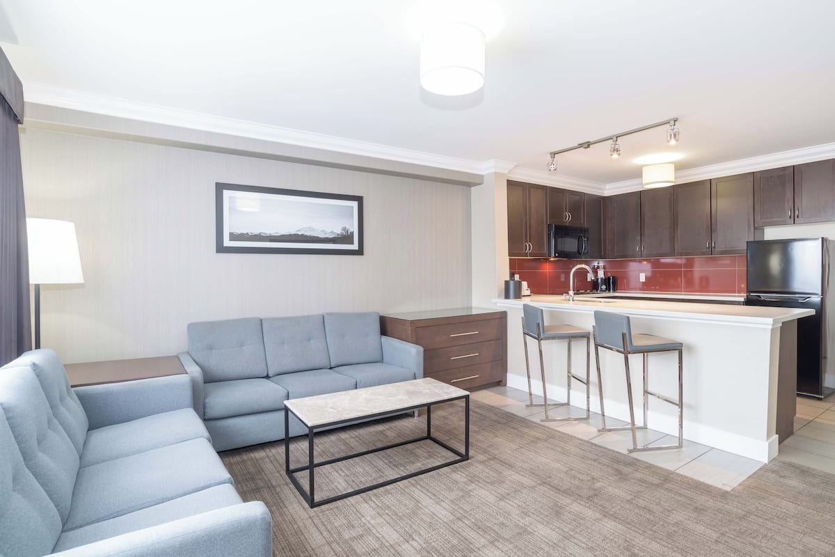 A modern living room with a grey sofa, a coffee table, and a kitchen area with a bar counter and stools.