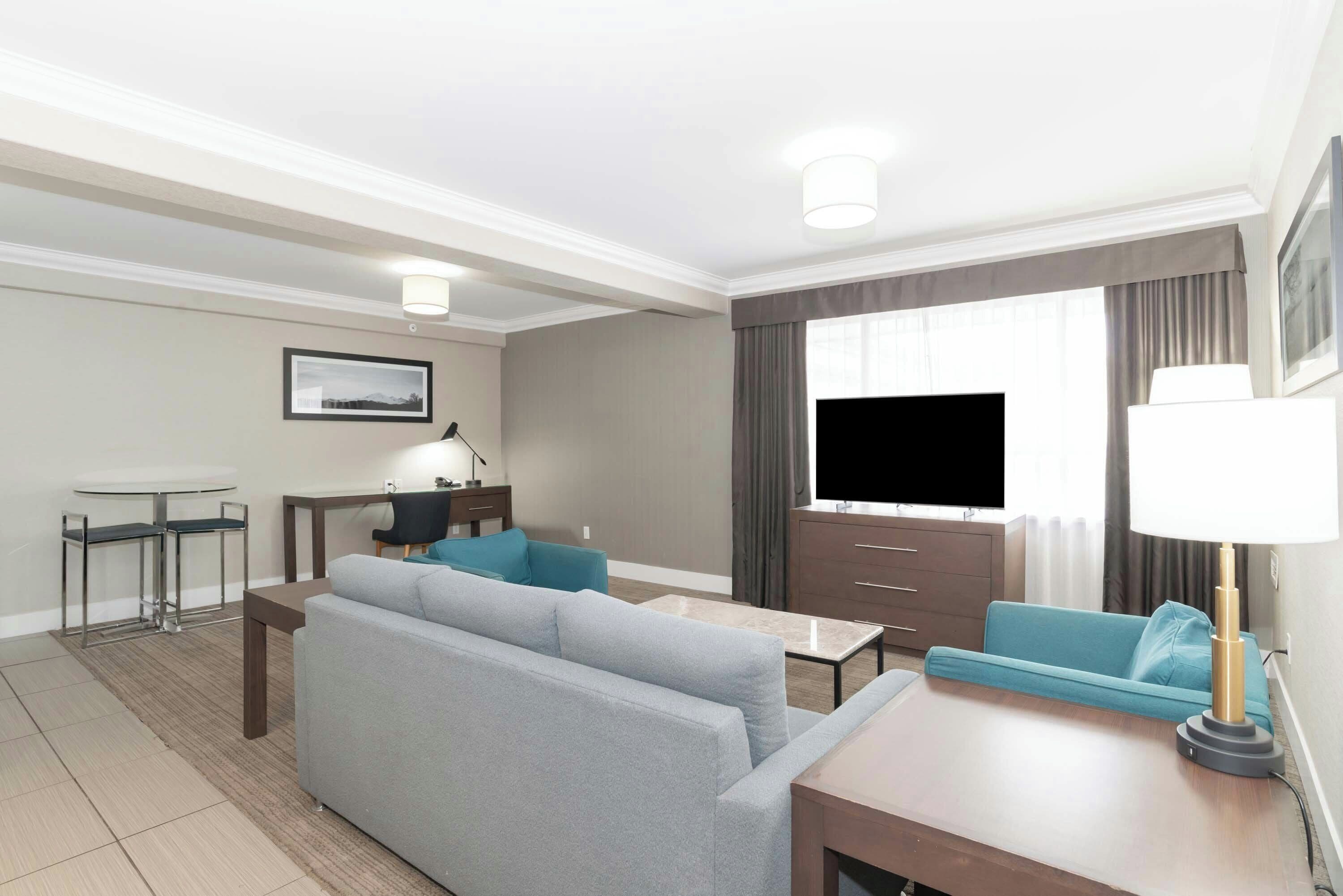A modern hotel room with a grey sofa, a small table, a TV, and a lamp.