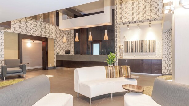A spacious living area with a white sofa, a grey armchair, and a wooden coffee table. A reception desk with a computer and a potted plant is visible in the background. - Sandman Hotel & Suites Abbotsford (Abbotsford)