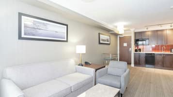 A hotel room with a sofa, a chair, a desk with a lamp, a picture frame, a kitchen area with cabinets and appliances, and a view of a body of water.