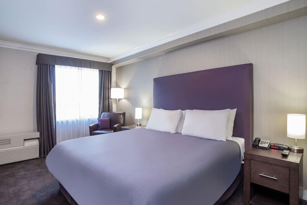 Ein Hotelzimmer mit einem großen Bett, einem Sessel, einem Schreibtisch mit Lampe, einem Telefon und einem Heizkörper.