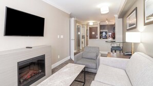 A modern living room with a flat-screen TV, fireplace, grey sofa, and a dining area with a table and chairs. - Sandman Hotel & Suites Abbotsford (Abbotsford)