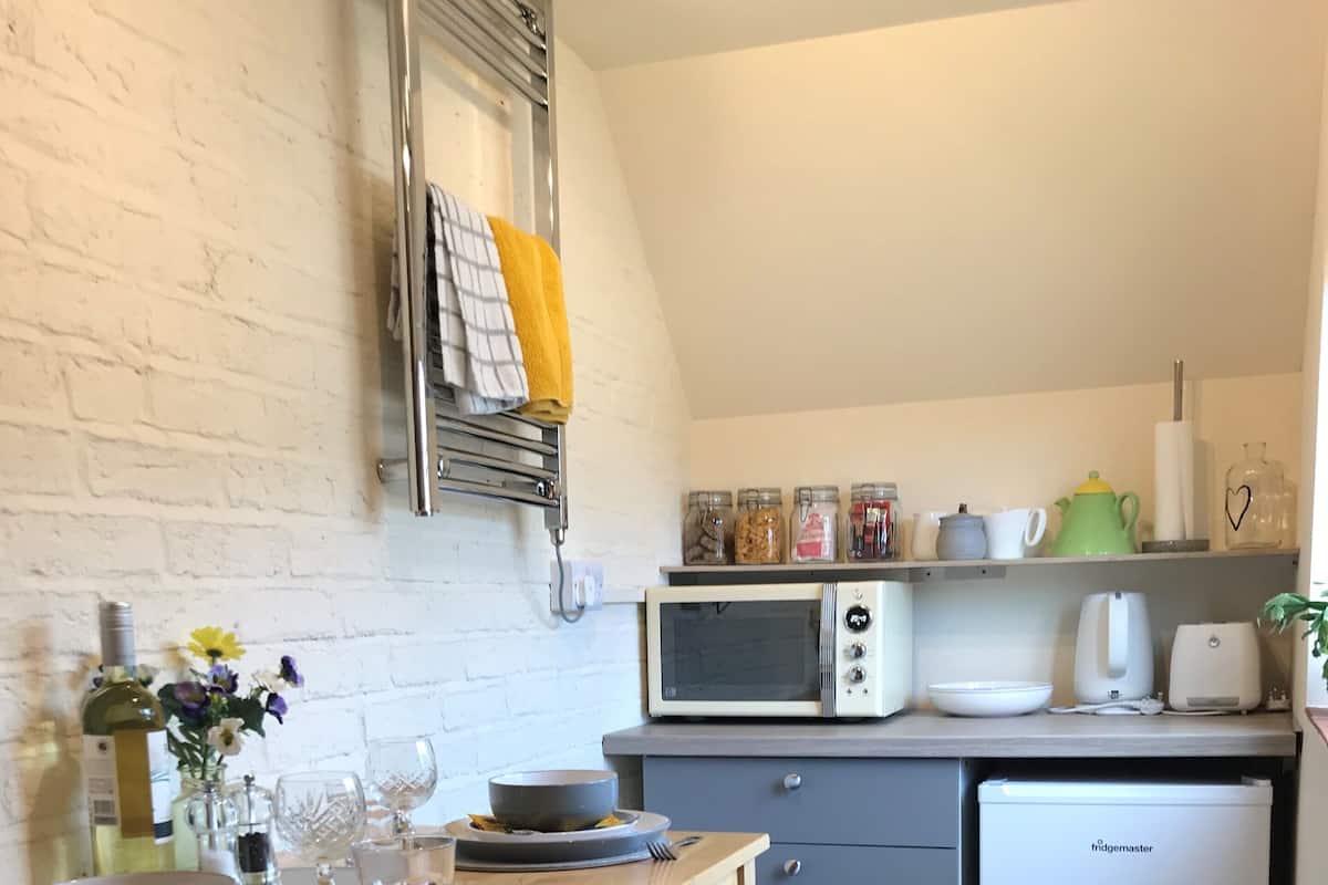 A compact kitchen with a white brick wall, a wooden table with a bowl and glasses, a microwave, and a fridge.