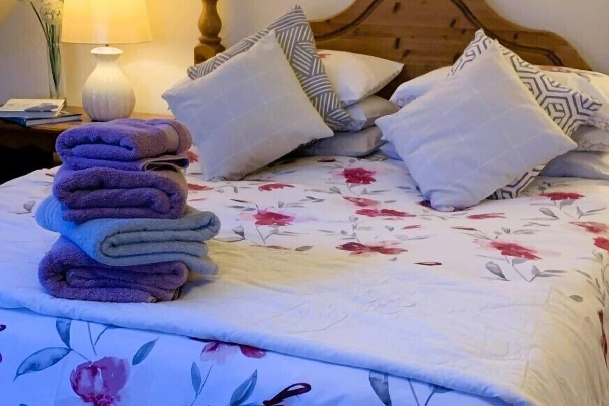 A neatly made bed with a floral bedspread, a stack of folded towels, and a bedside lamp.
