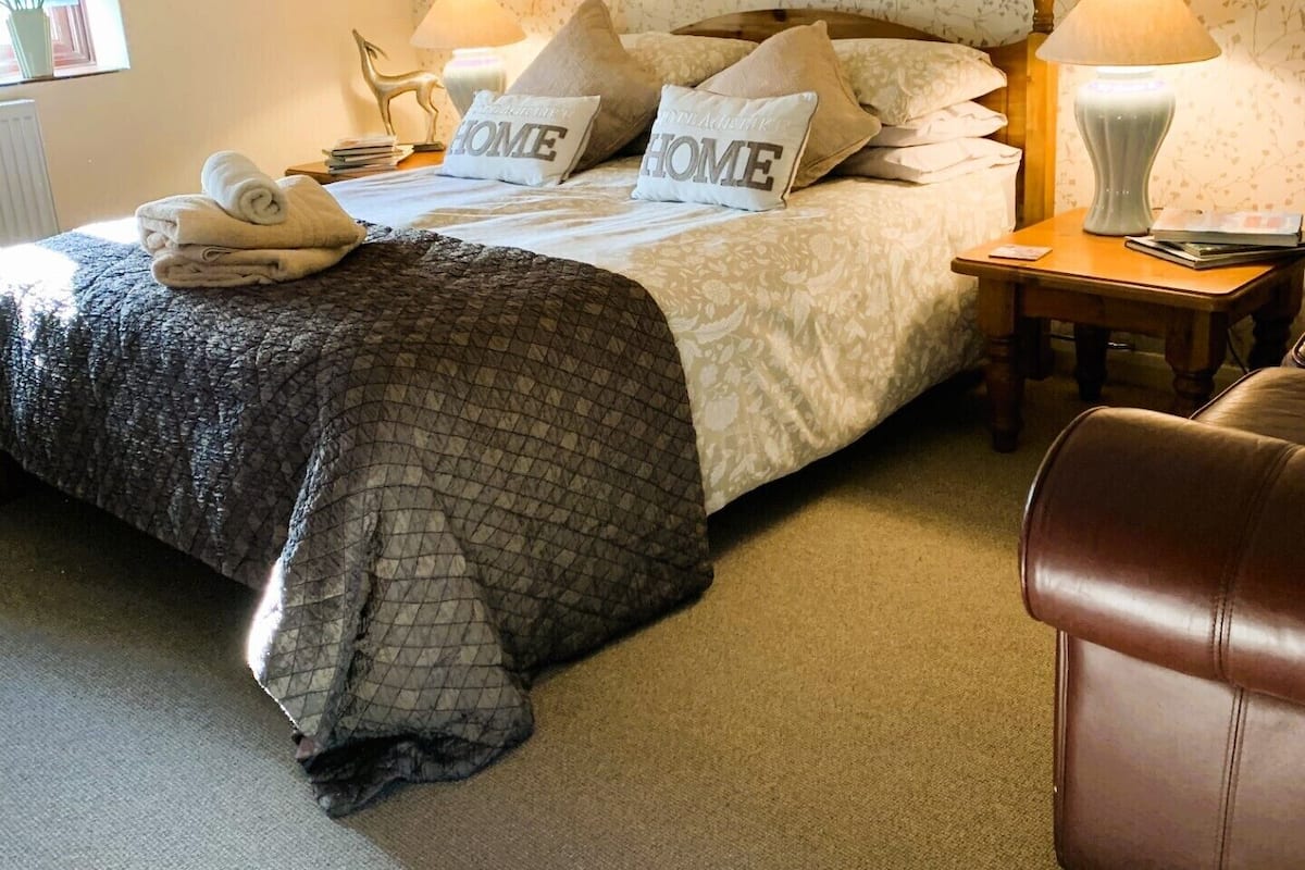 A neatly arranged bedroom with a bed, pillows, a bedside table with a lamp, a chair, and a window with flowers.