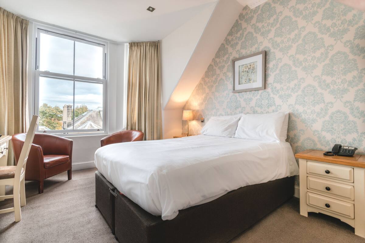 A bedroom with a large bed, a desk, a chair, a nightstand, and a window with curtains.