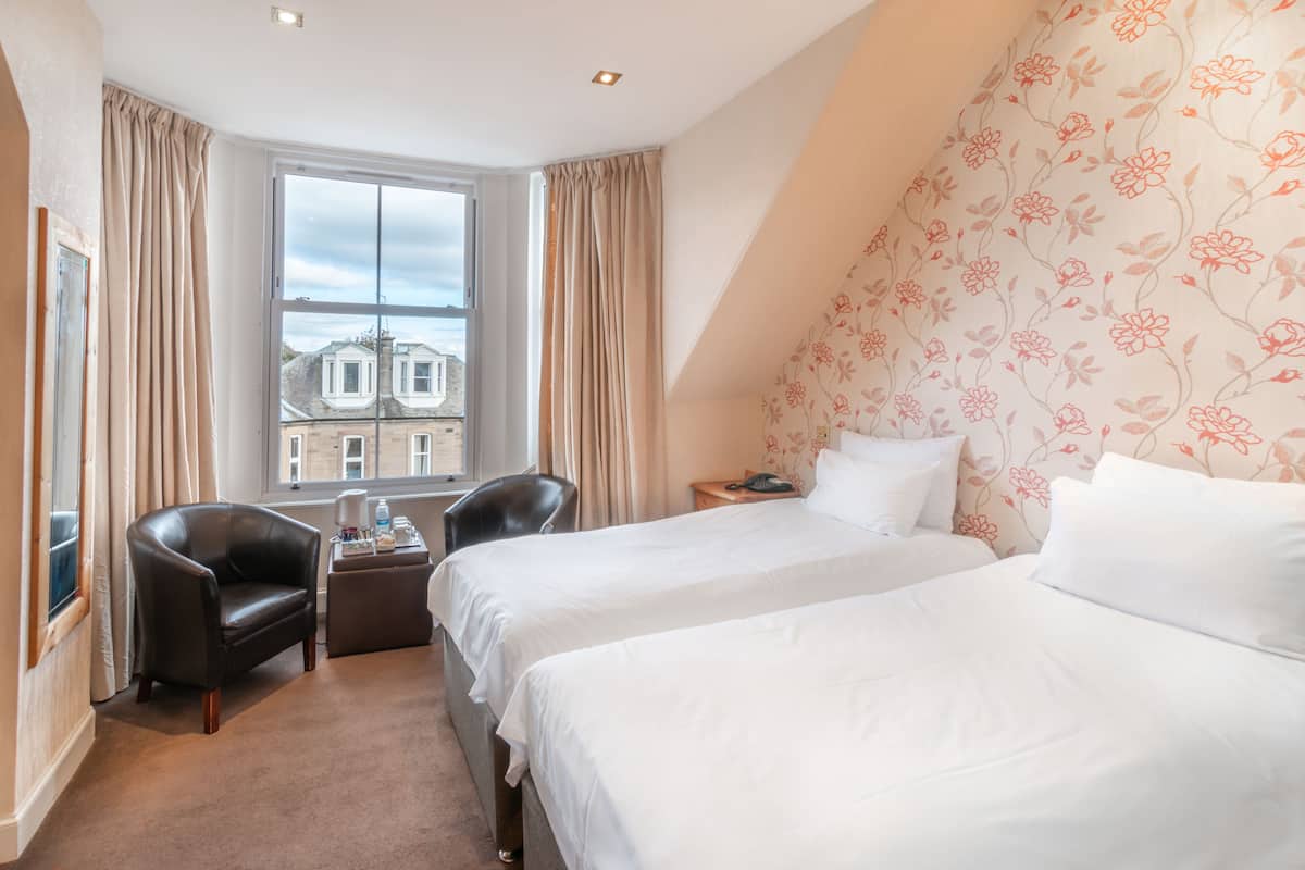 A hotel room with two beds, a chair, a small table with a phone, and a window with a view of buildings.
