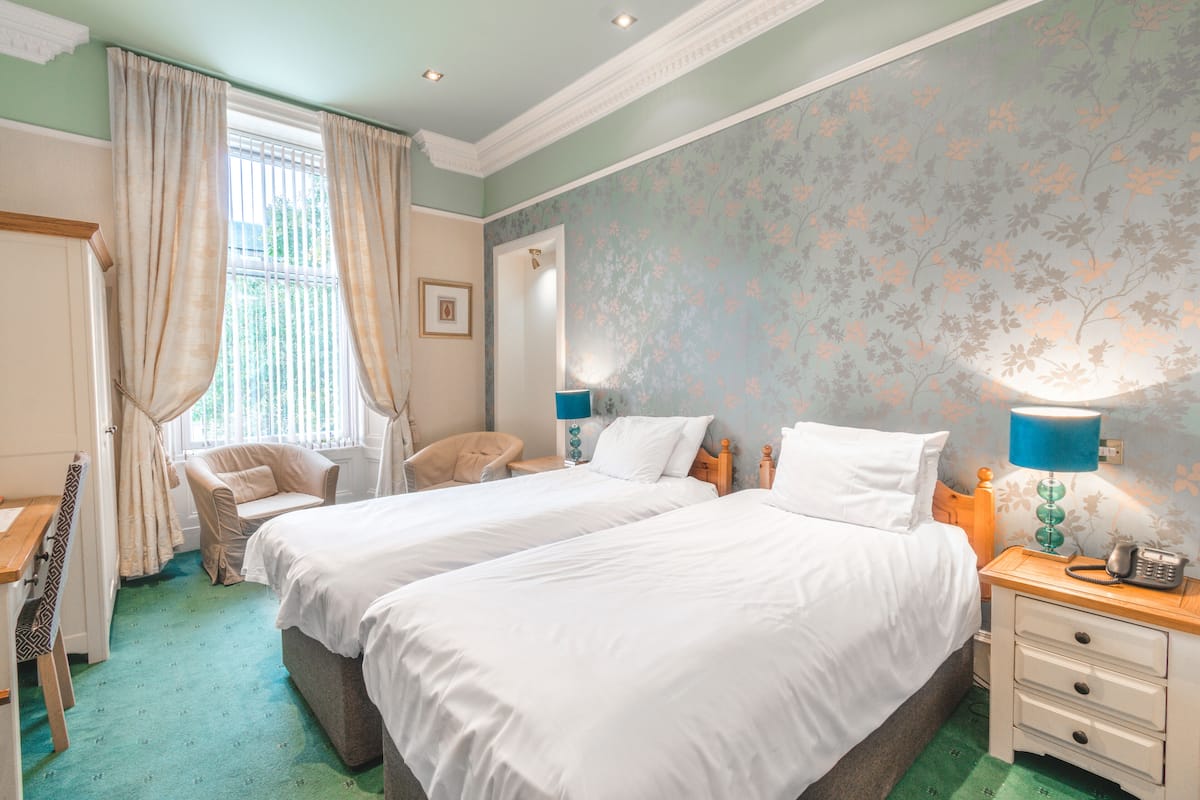 A hotel room with two beds, a desk, a chair, and a nightstand with a lamp.