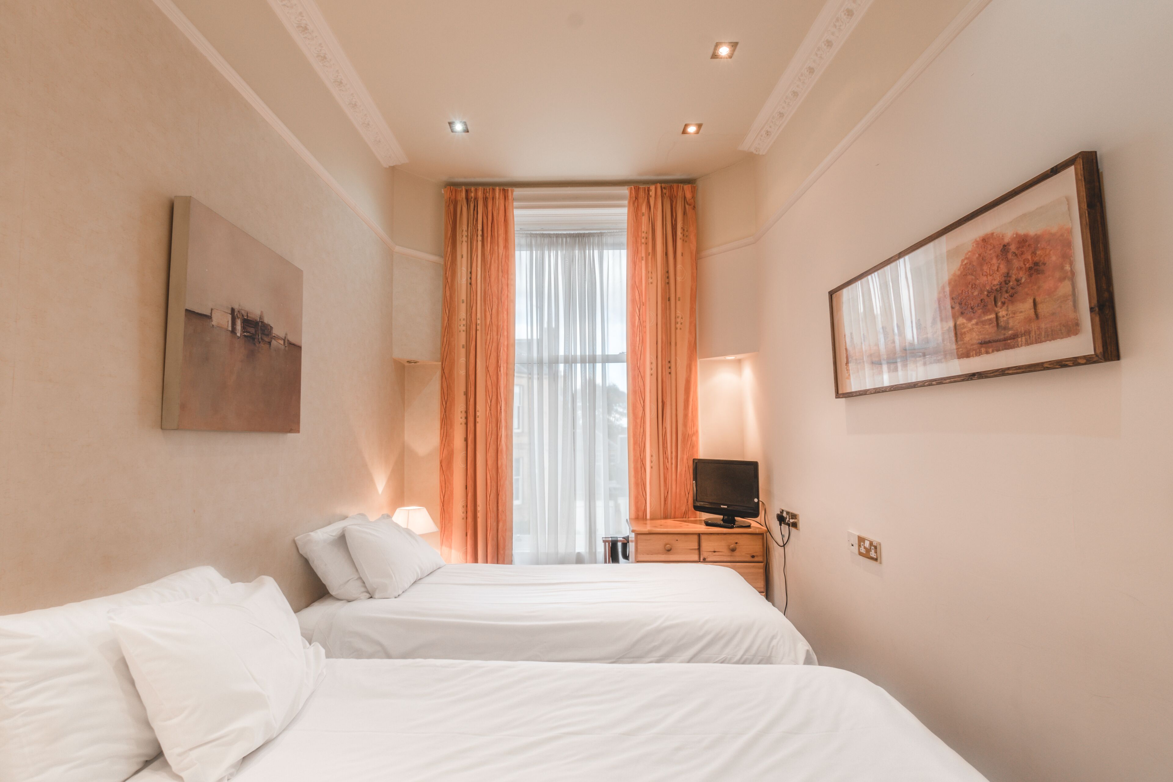 Una habitación de hotel con dos camas, un televisor, una ventana con cortinas y obras de arte en las paredes.