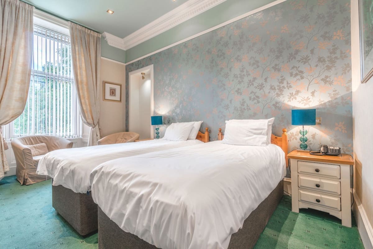 A double bed with white linens, a bedside table with a lamp, a chair, and a floral wallpapered wall.