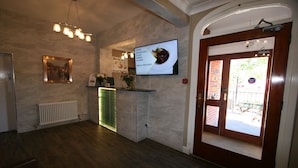 A reception area with a wooden floor, a reception desk, a wall-mounted TV displaying a menu, and a door leading outside. - Grange Moor Hotel (Maidstone)