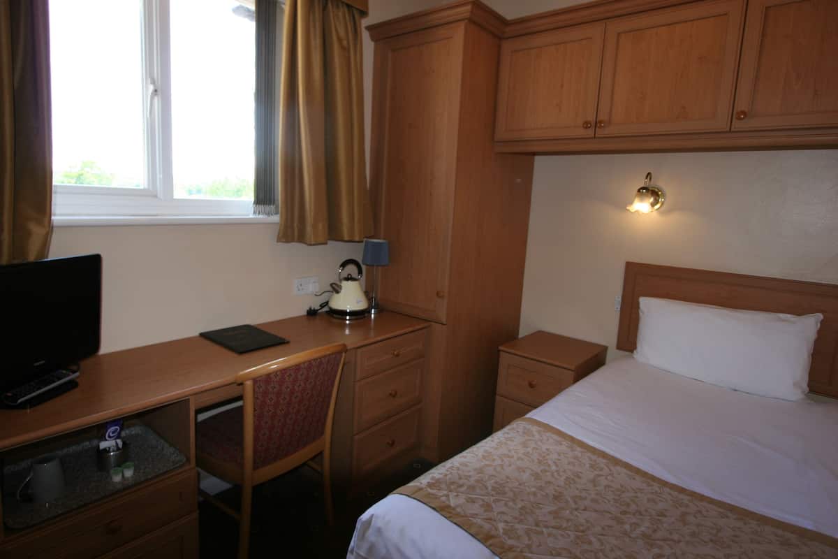 Ein Hotelzimmer mit Schreibtisch aus Holz, einem Fernseher, einem Bett mit weißem Kopfteil und einem Nachttisch mit Lampe.