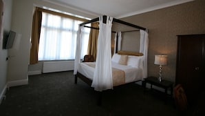 A four-poster bed with white linens, a bedside table with a lamp, and a view of the outdoors through large windows. - Grange Moor Hotel (Maidstone)