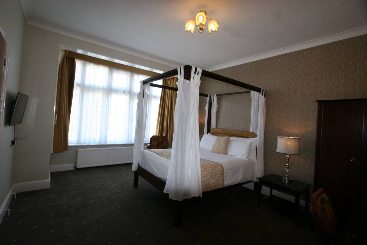 Ein Bett mit Baldachin, ausgestattet mit weißer Bettwäsche, ein Nachttisch mit Lampe und Blick ins Freie durch große Fenster.