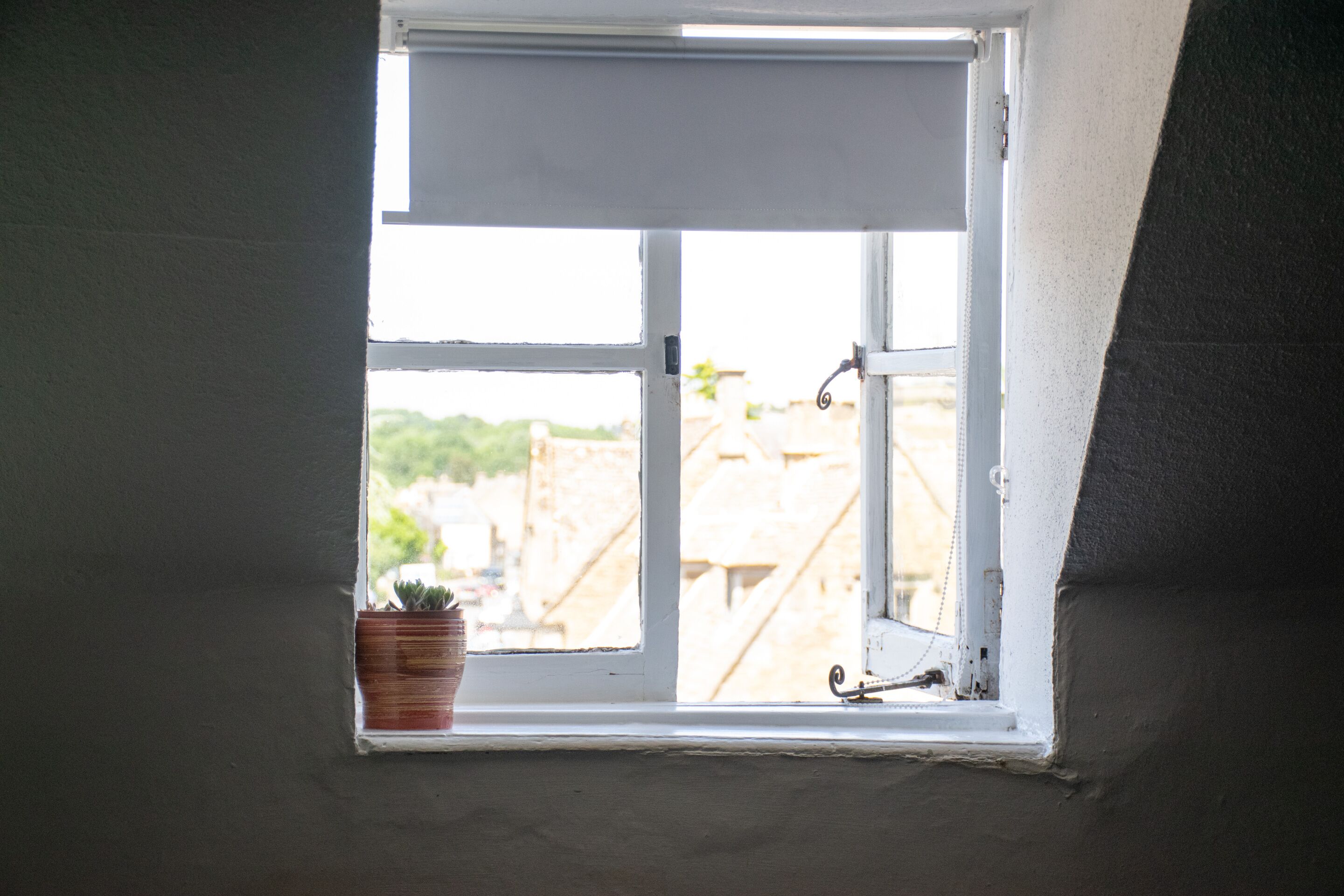 Ein kleines Fenster mit weißem Rollo, einer Topfpflanze und einem Metallriegel.