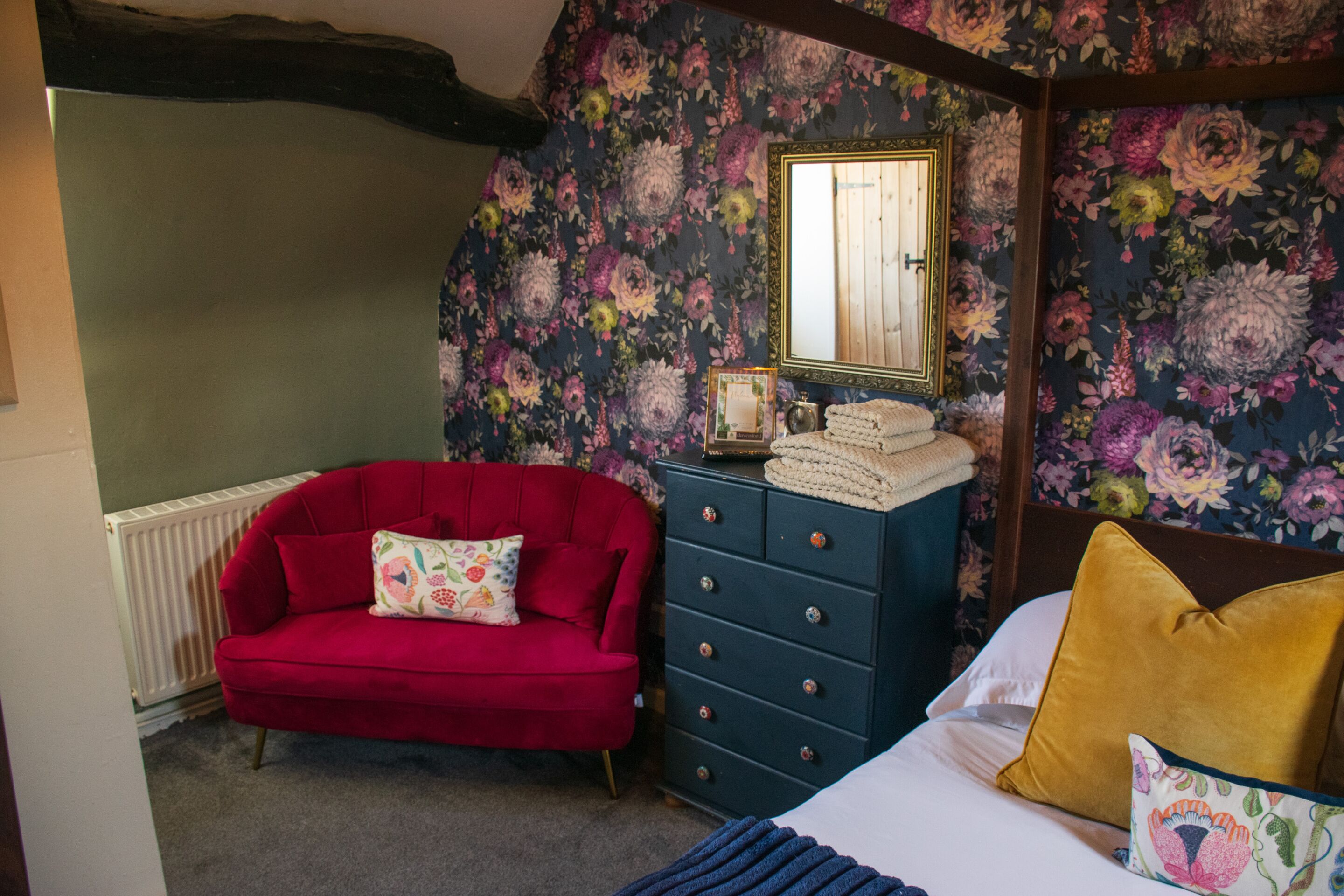 A bedroom with a red sofa, a dresser, a bed with a yellow pillow, and floral wallpaper.