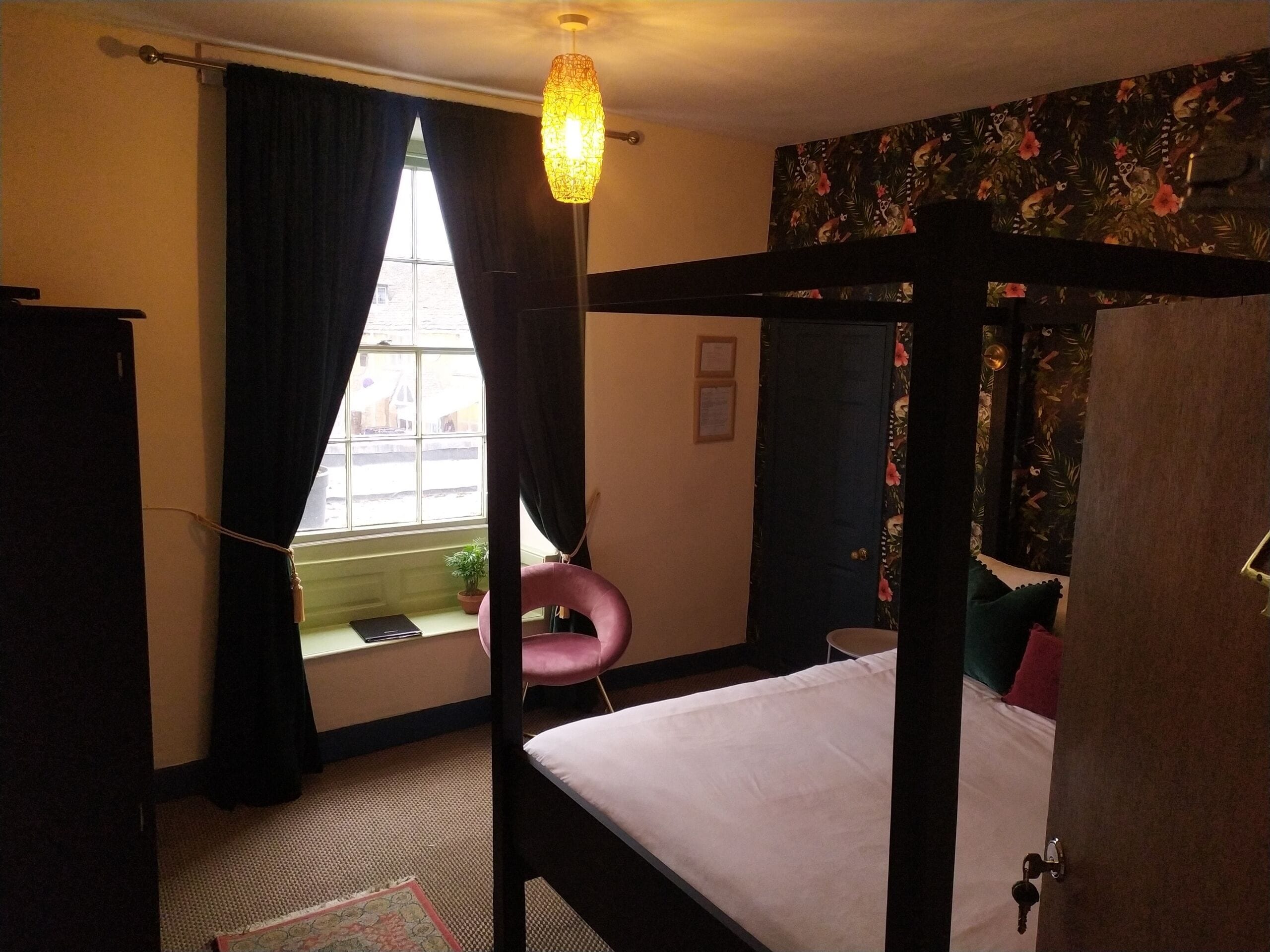 A room with a four-poster bed, a window with curtains, a potted plant, and a floral wallpaper.