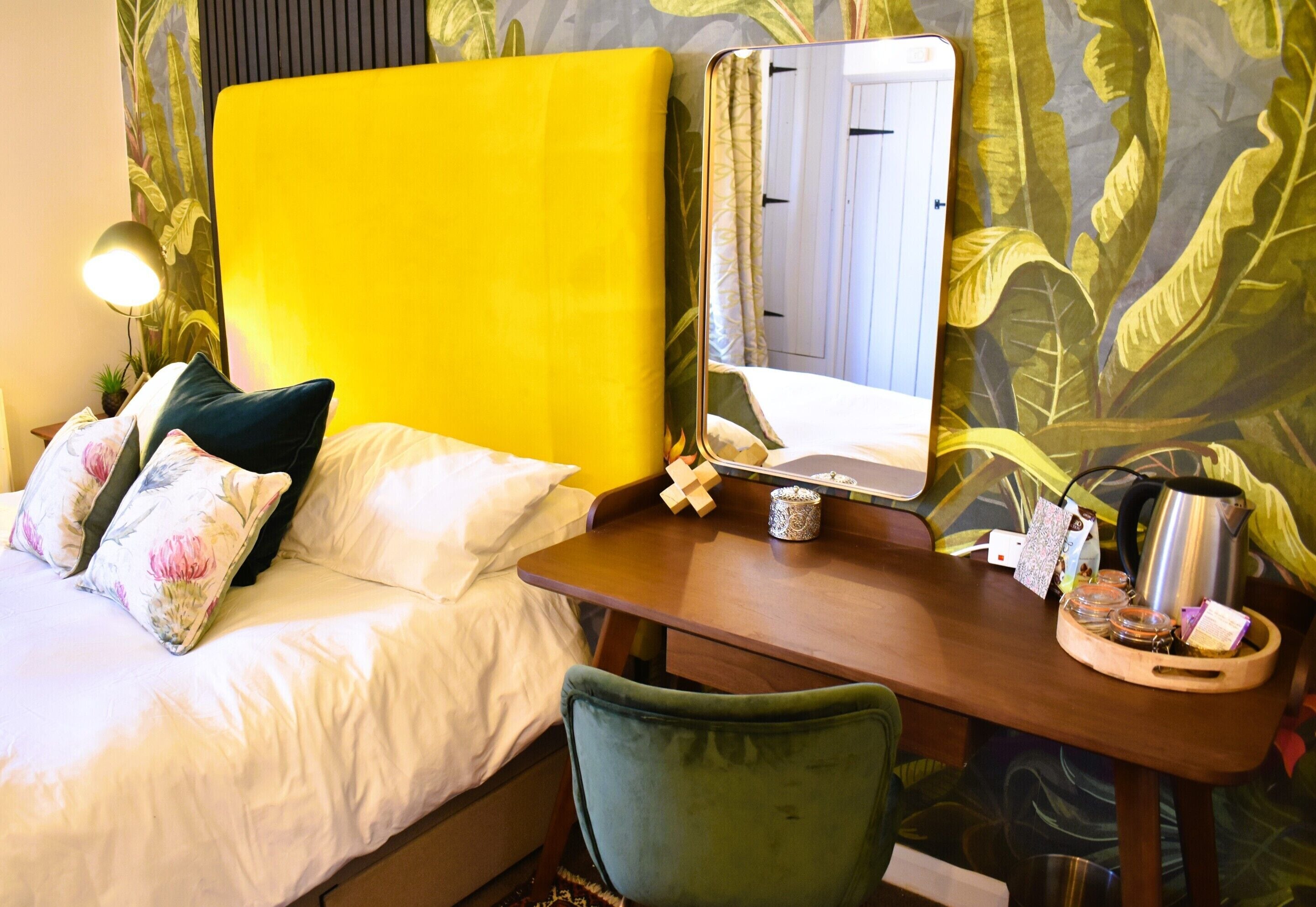 Habitación de hotel con cabecera amarilla, cama con almohadas blancas y florales, una silla verde, un escritorio de madera con un espejo, una tetera y una bandeja con frascos.