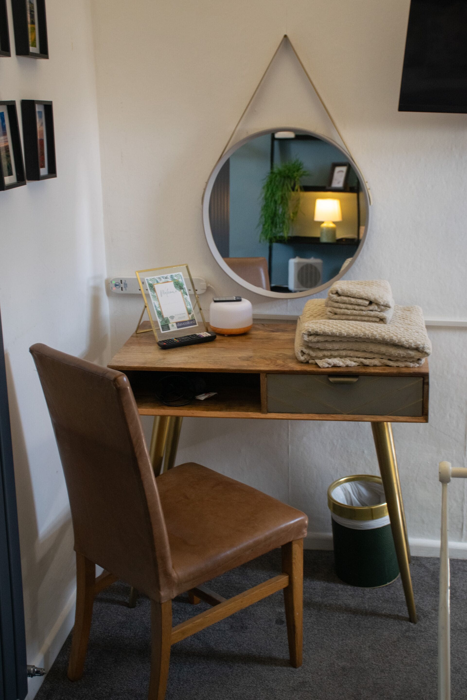 A hotel room with a wooden desk, a brown leather chair, a round mirror, a lamp, a picture frame, a small trash bin, and a stack of towels.