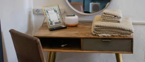 A hotel room with a wooden desk, a brown leather chair, a round mirror, a lamp, a picture frame, a small trash bin, and a stack of towels.