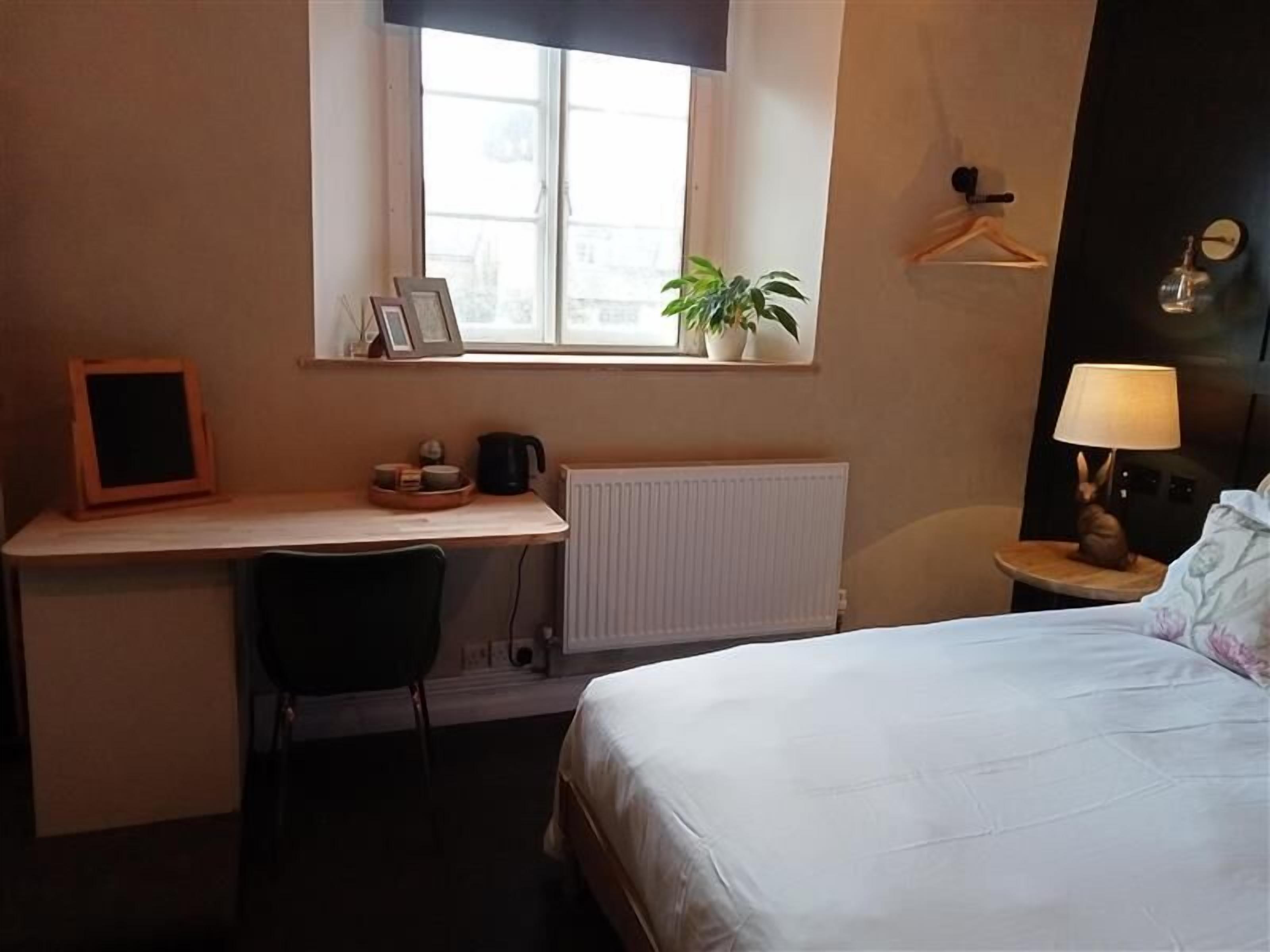 A hotel room with a bed, a desk with a chair, a radiator, a window with a plant, and a lamp on a bedside table.