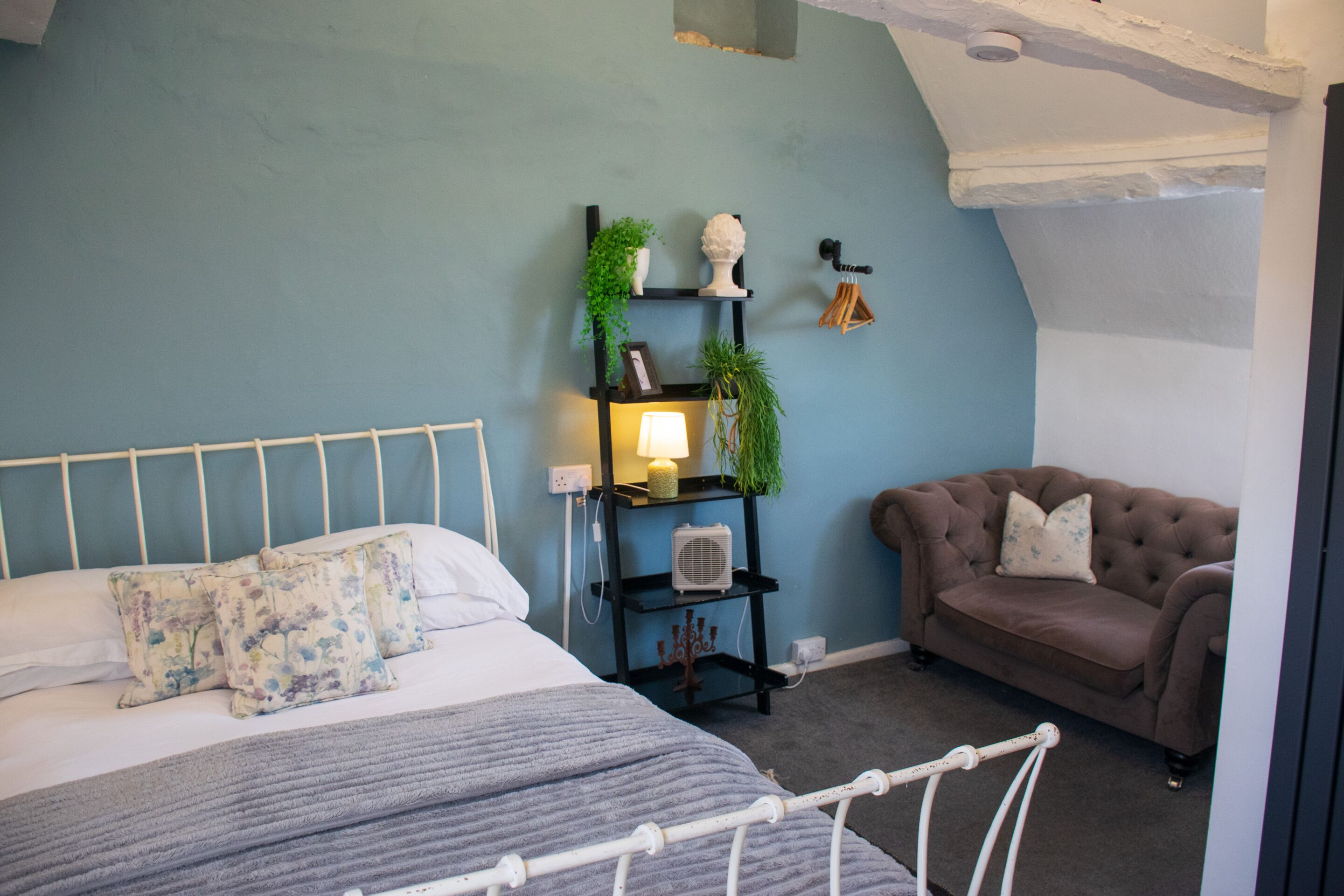 A bedroom with a bed, a bedside table with a lamp, a small shelf with plants, and a brown armchair.