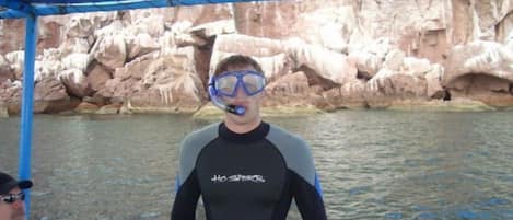 A person in a black and grey wetsuit sitting on a boat with a rocky shoreline in the background.