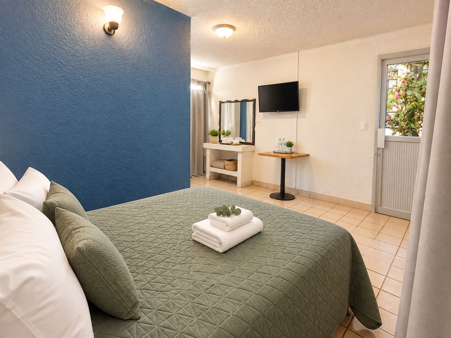 A hotel room with a bed, a bedside table with a mirror, a TV mounted on the wall, and a window with a view of greenery.