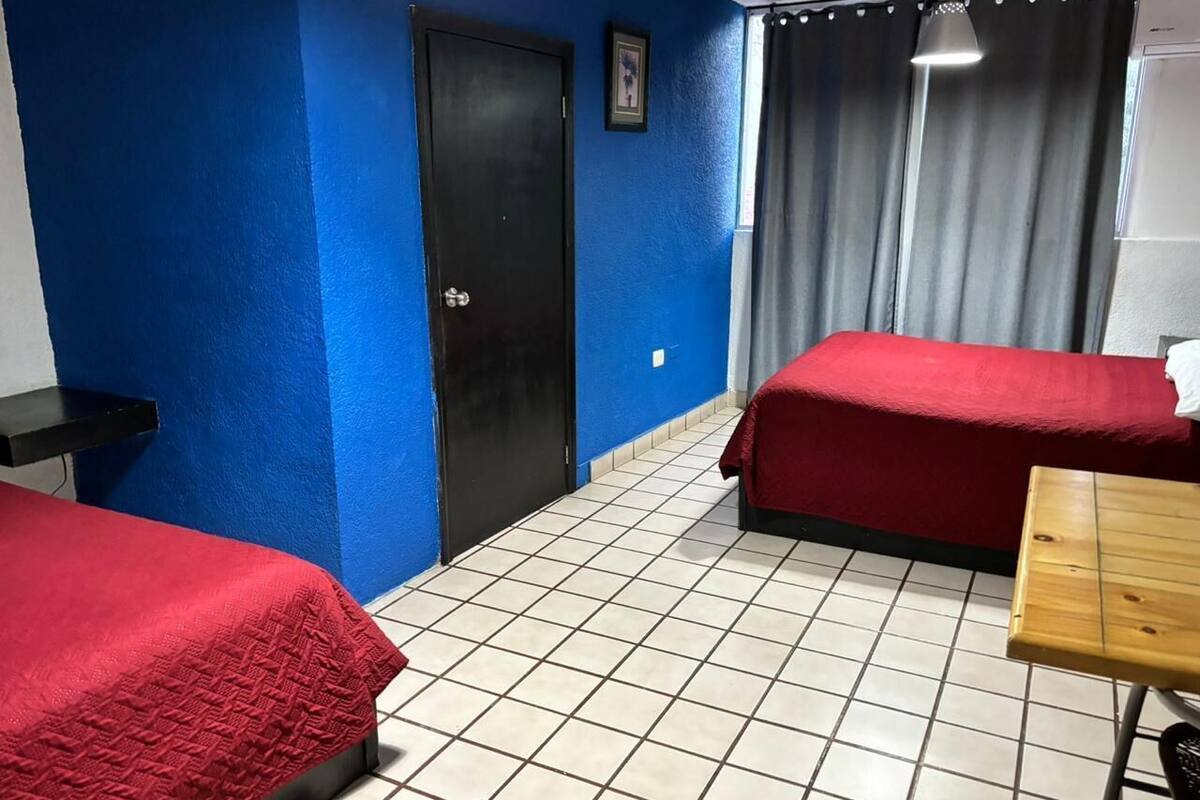 Una habitación con dos camas, una mesa de madera, una puerta negra y una ventana con cortinas.