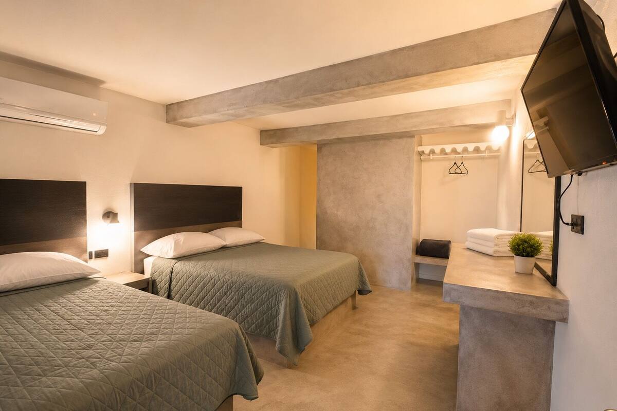 Habitación de hotel con dos camas, televisión, baño pequeño y una pared de acento de concreto.