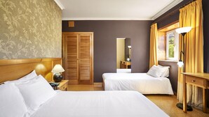A hotel room with two beds, a wooden wardrobe, a desk with a lamp, and a window with curtains.