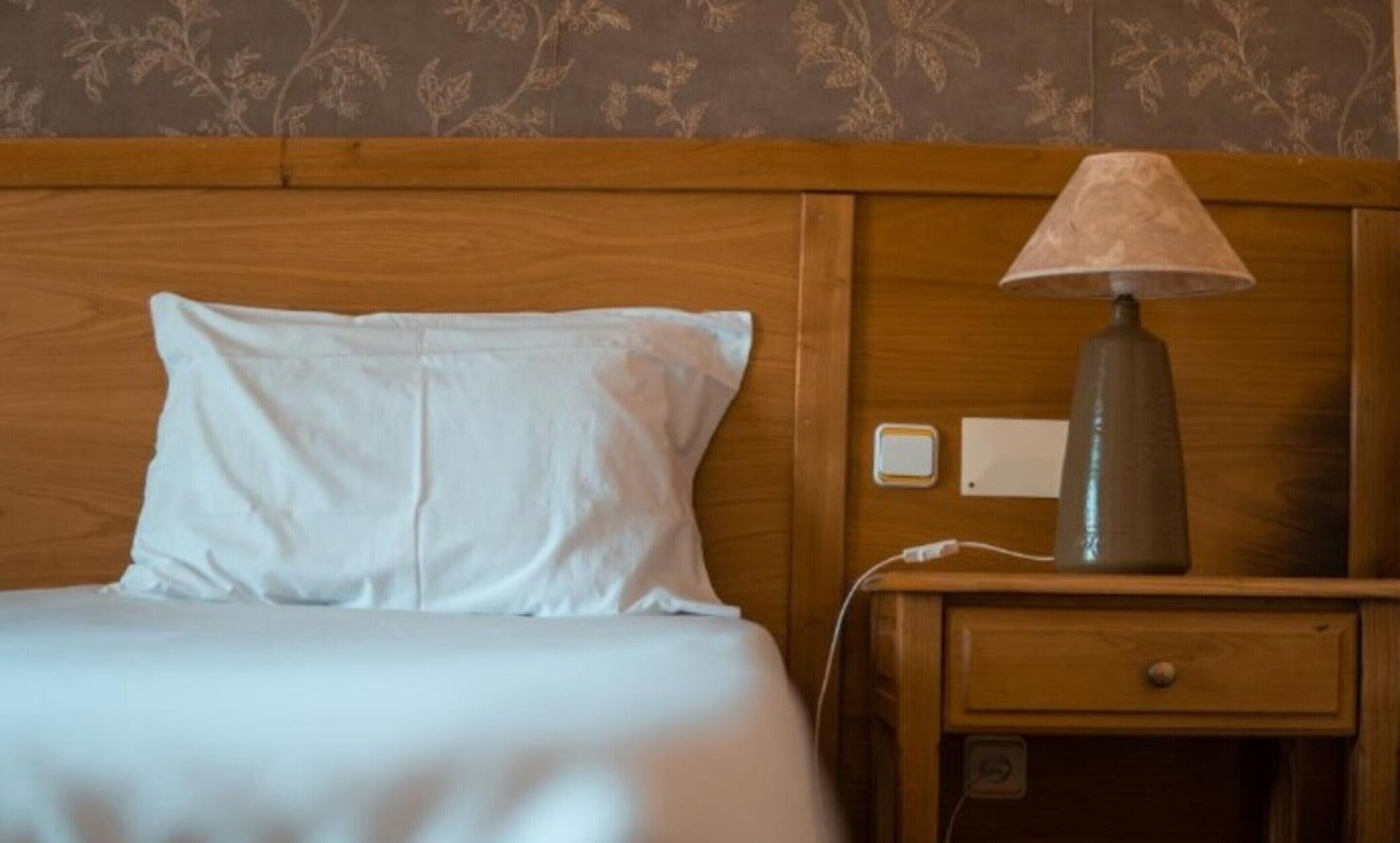A neatly made bed with a white pillow and a wooden nightstand with a lamp.