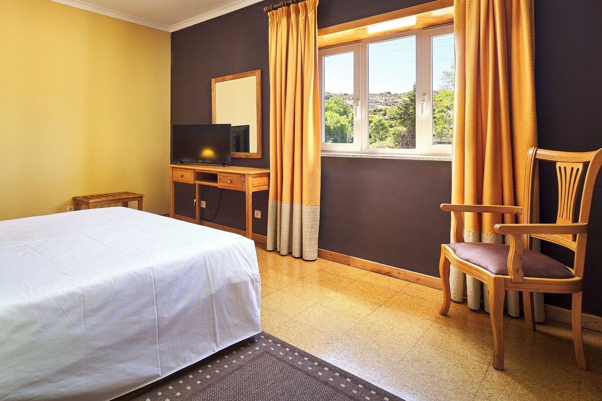A hotel room with a bed, a desk with a computer, a chair, and a window with curtains.