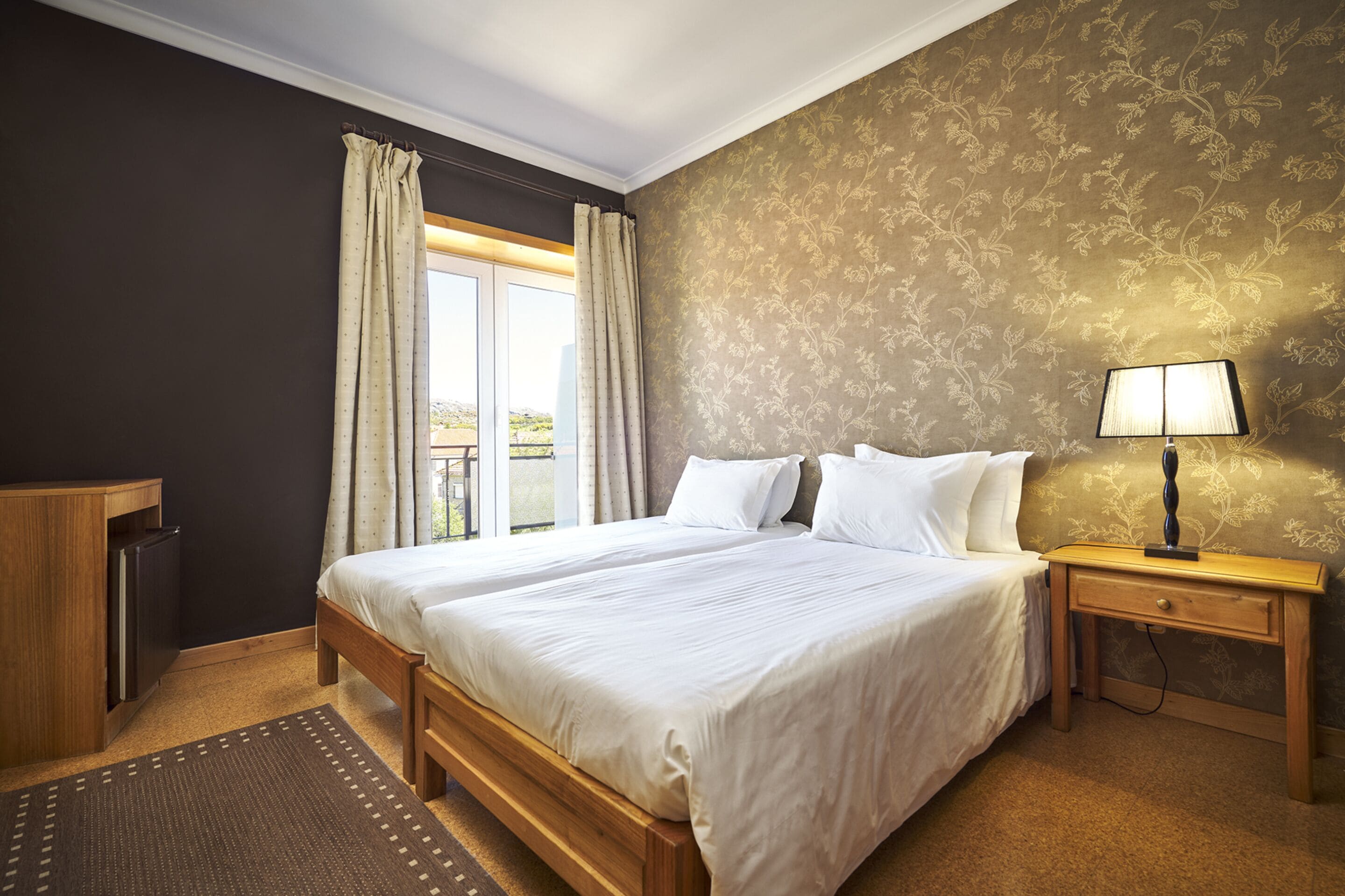 A neatly arranged bedroom with a large bed, a bedside table with a lamp, a wardrobe, and a window with curtains.