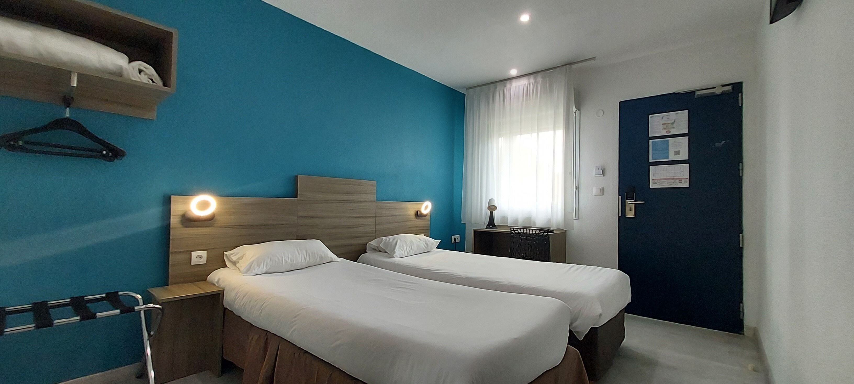 Een hotelkamer met twee bedden, een blauwe accentmuur, een houten hoofdbord, een kapstok en een deur met papieren erop.