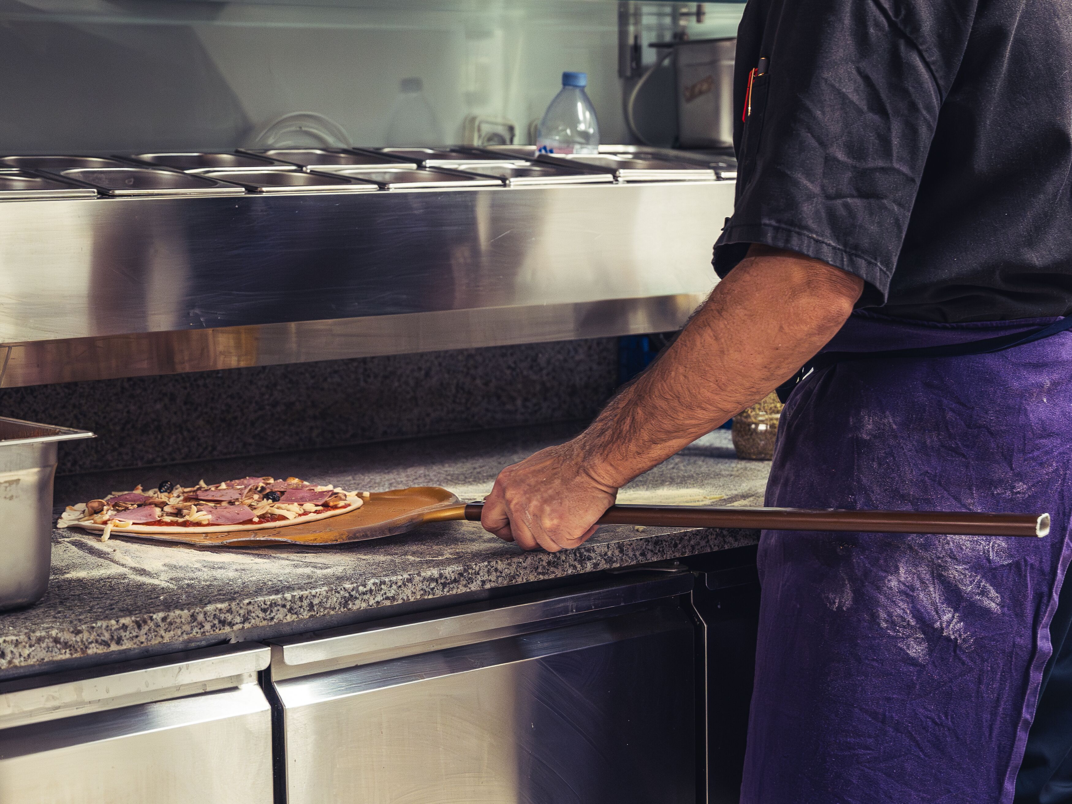 A kitchen with a pizza on a wooden board, a person in a dark shirt and purple apron, and stainless steel appliances.