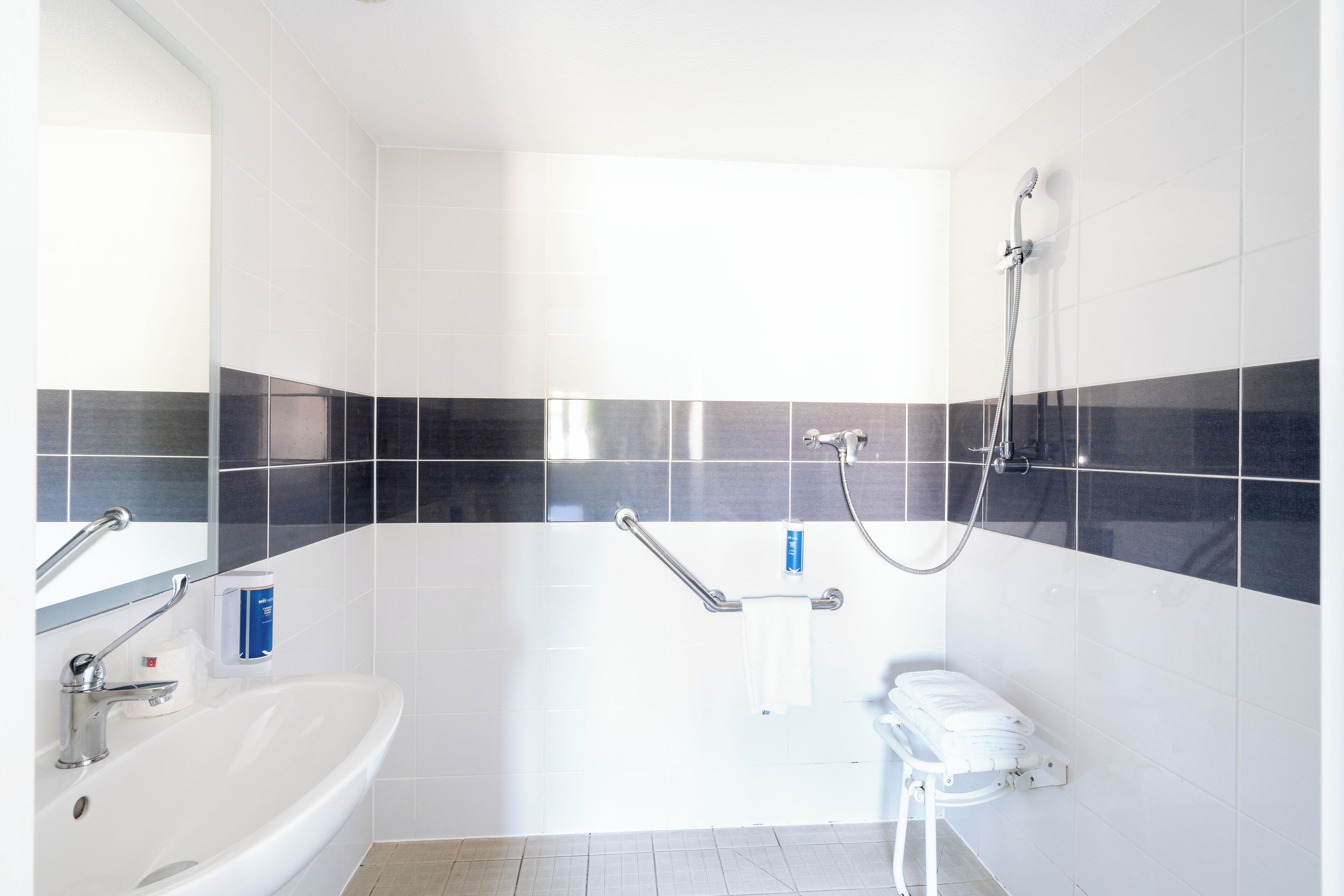 Un baño moderno con un lavamanos blanco, un espejo, un cabezal de ducha de mano y un inodoro con barra de apoyo.