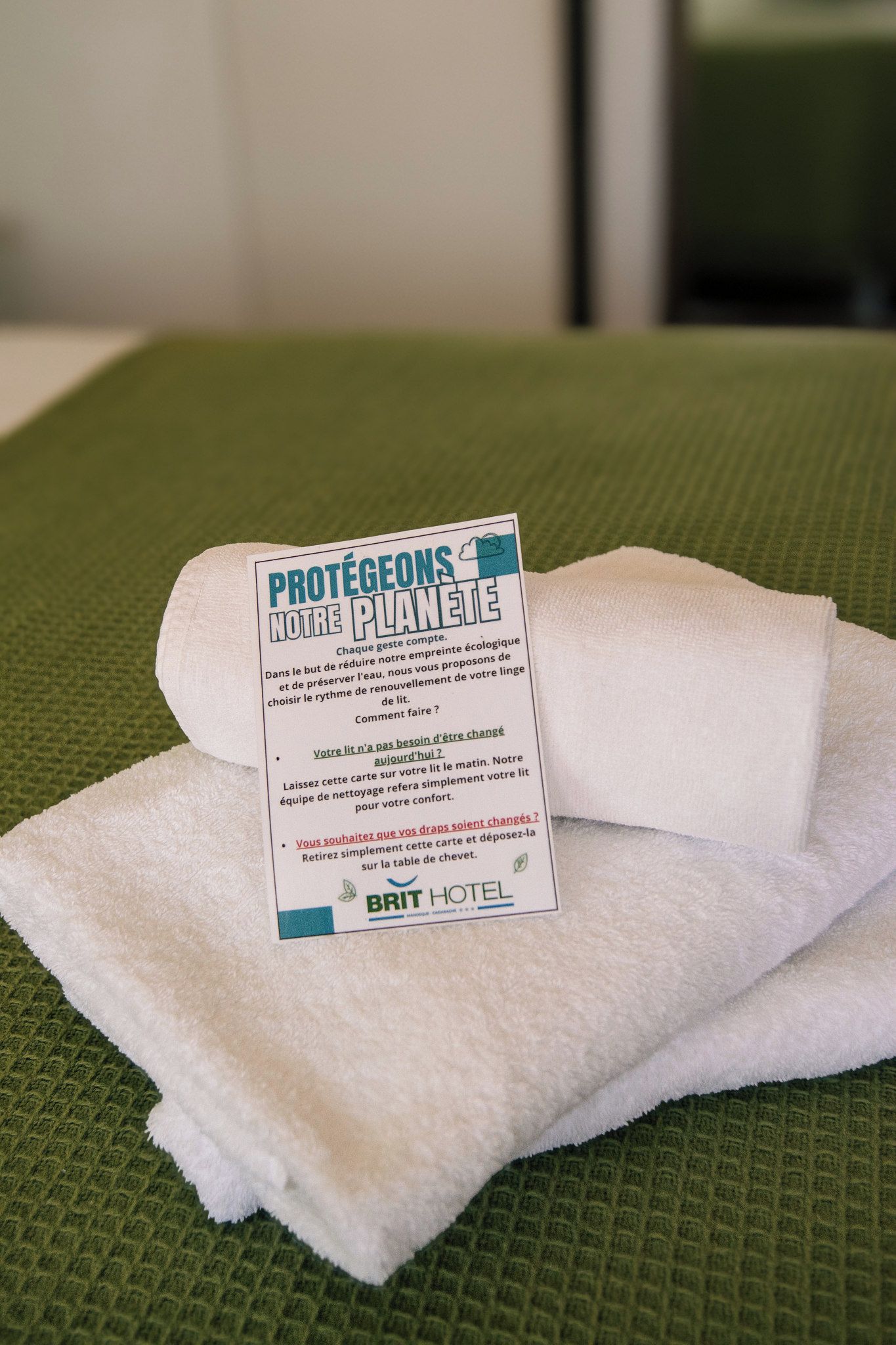 Una toalla blanca con un mensaje impreso en francés sobre una superficie verde texturizada, probablemente un felpudo de baño o calentador de toallas.