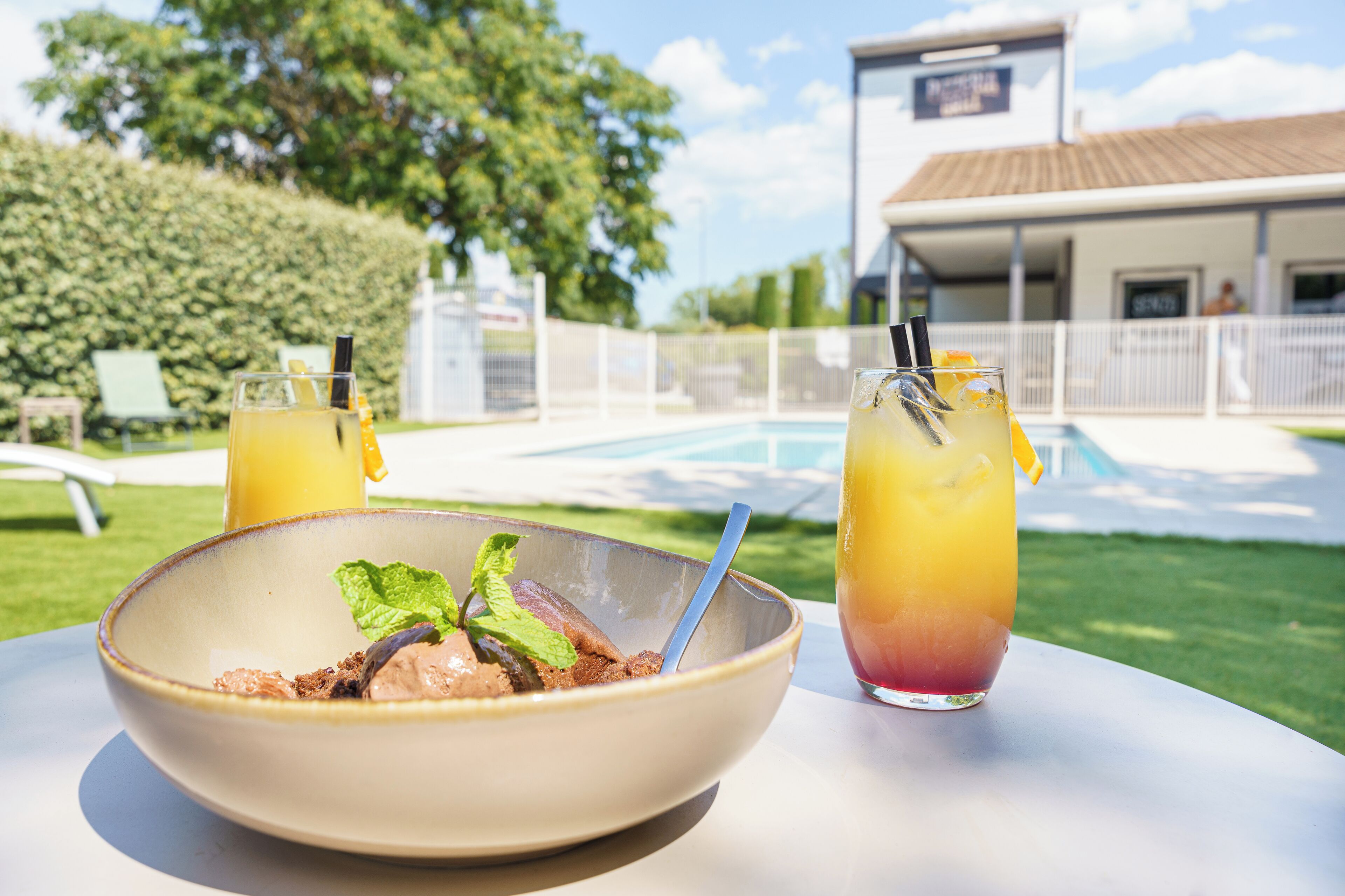 Une table à manger avec un saladier de dessert et deux boissons, une piscine et un bâtiment en arrière-plan.
