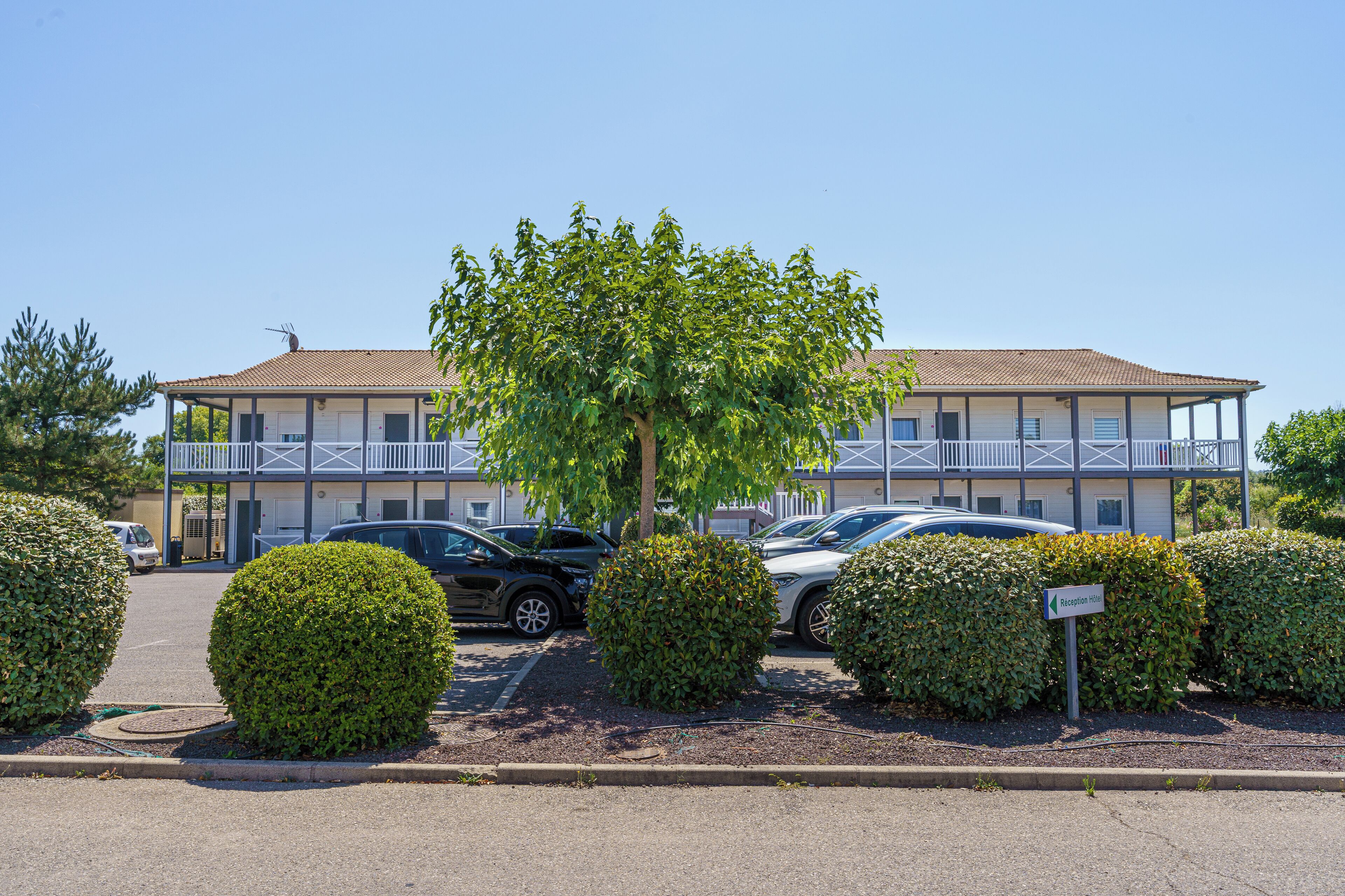 A two-story motel with a balcony on the second floor, a parking lot with several cars, and well-maintained landscaping.