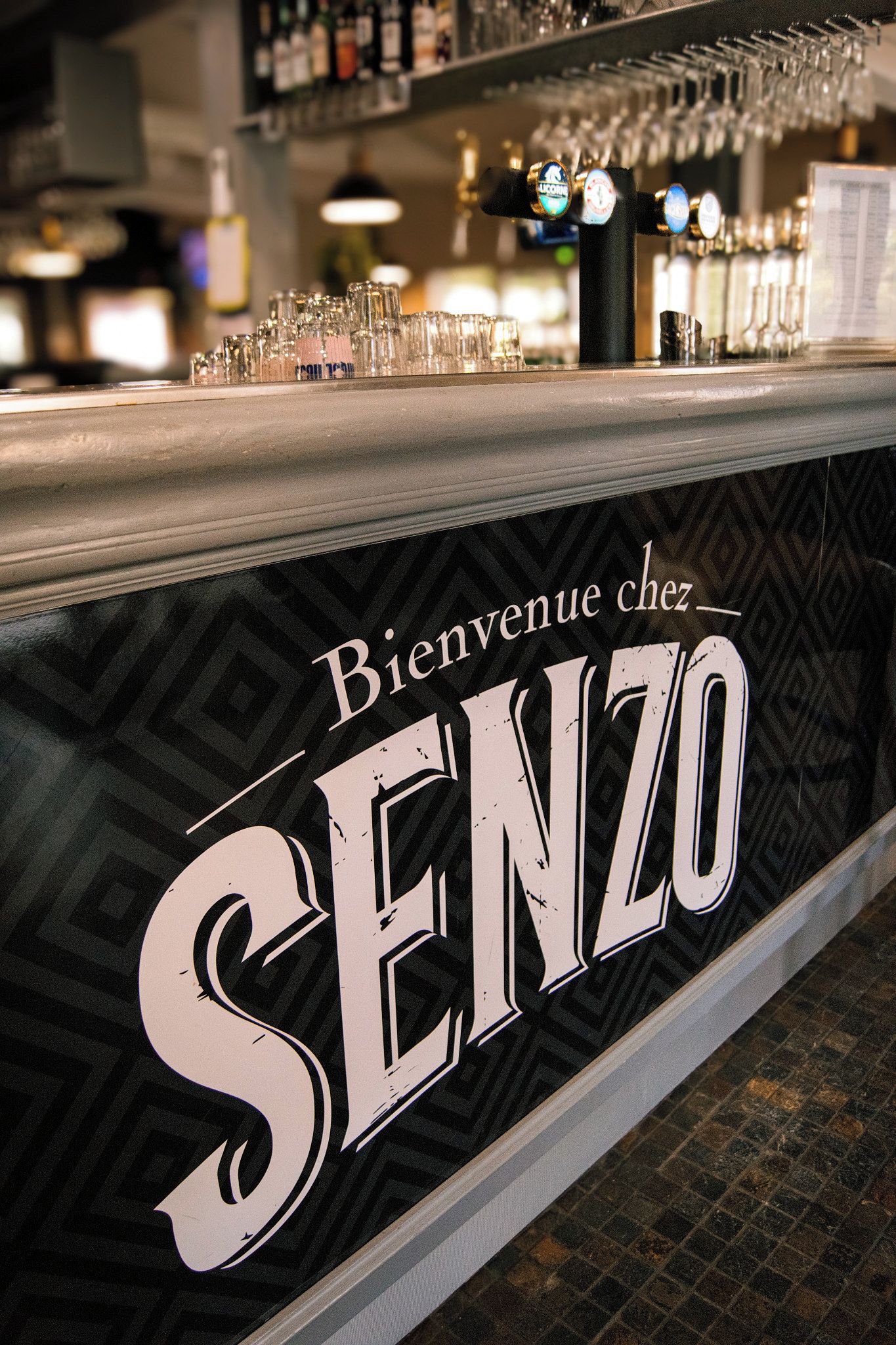 Una barra con un cartel que dice 'Bienvenue chez SENZO' y una selección de vasos y botellas.
