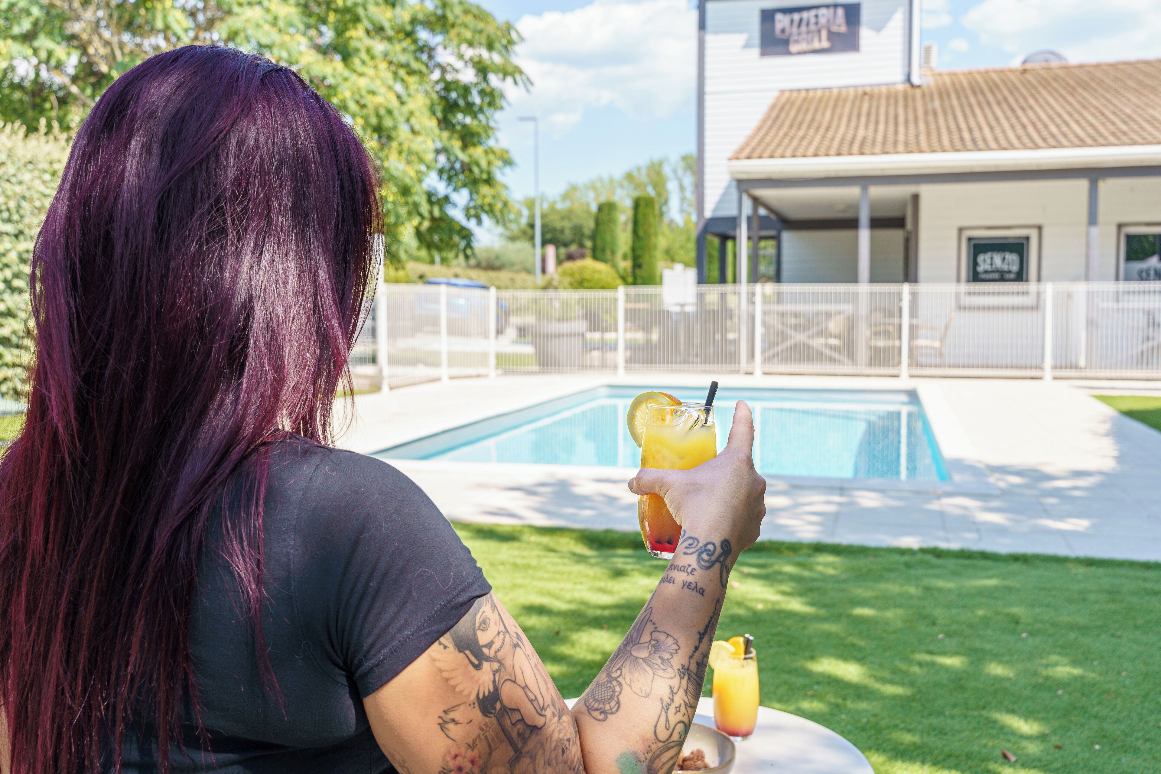 Eine Person mit lila Haaren sitzt an einem Tisch mit einem Getränk und blickt auf einen Swimmingpool und ein Gebäude mit einem Schild.