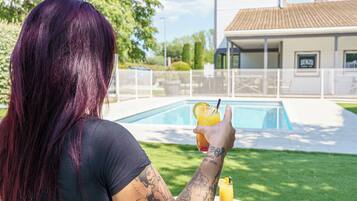 Eine Person mit lila Haaren sitzt an einem Tisch mit einem Getränk und blickt auf einen Swimmingpool und ein Gebäude mit einem Schild.