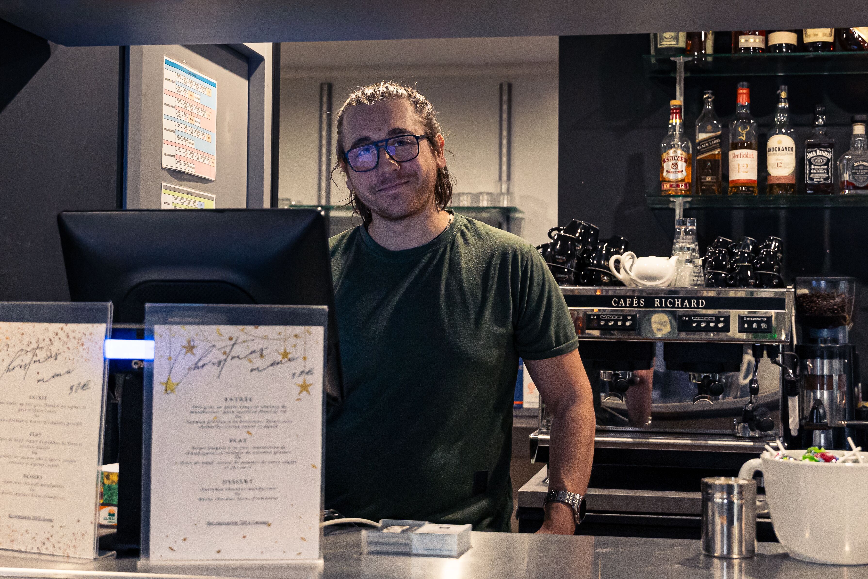 A barista at a counter with a menu, espresso machine, and a selection of liquor bottles in the background.