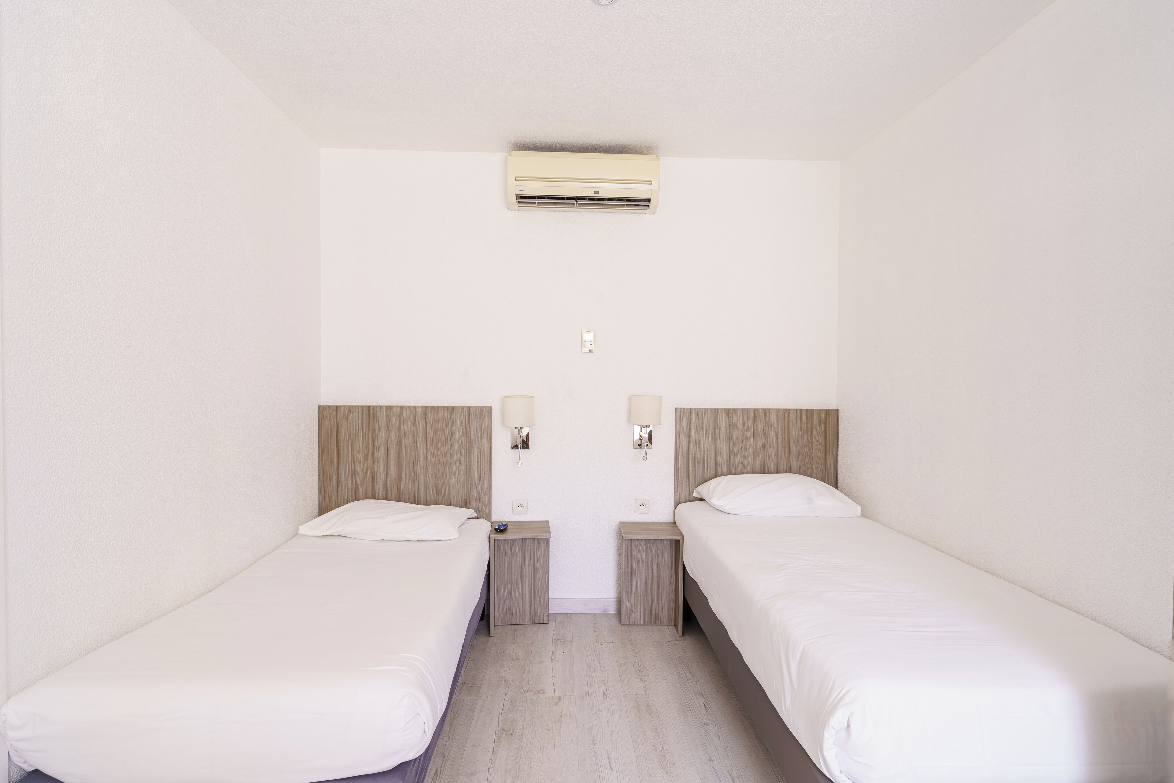 Habitación de hotel con dos camas individuales, una mesita de noche pequeña con lámpara, un equipo de aire acondicionado en la pared y un enchufe.
